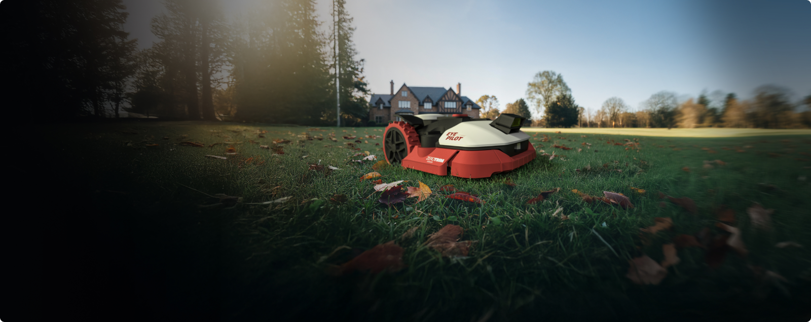 Kress EyePilot robotic mower mowing a leaf-strewn lawn at sunrise in front of a large house